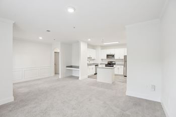 A large, empty, white room with a carpeted floor and a kitchen in the background.
